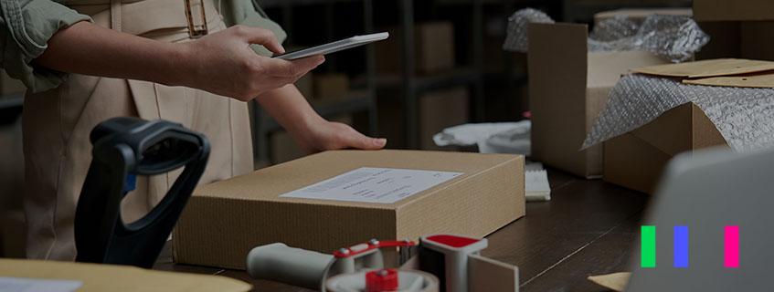 Mão segurando um celular e apontando a câmera dele para o qr code de uma caixa de papelão que estão sobre a mesa.