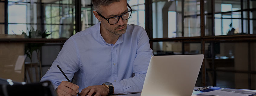 Homem de meia idade sentando em frente a uma mesa de escritório, olhando para a tela do notebook e escrevendo em uma papel - PDV Digital