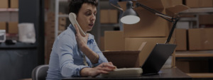 Mulher sentada em frente para uma mesa mexendo no seu notebook, dentro um galpão e falando ao telefone