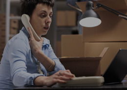 Mulher sentada em frente para uma mesa mexendo no seu notebook, dentro um galpão e falando ao telefone