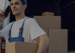 Homem segurando uma encomenda que está na caixa de papelão e ao fundo, uma van com várias caixas