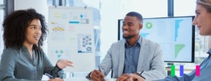 Três pessoas conversando em uma sala de reunião com gráficos no fundo.